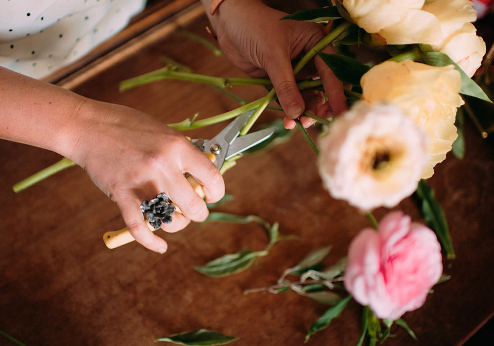 Hands trim flower stems with pruning shears on a wooden table. Several blooming flowers and scattered leaves surround the area.