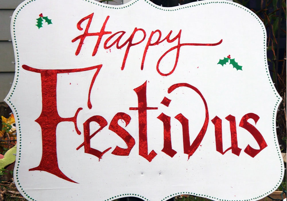 A white sign displaying "Happy Festivus" in red calligraphy, adorned with small holly decorations, is placed outdoors against a backdrop of a house wall and garden foliage.