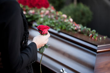 A hand holding a red rose beside a closed, wooden casket adorned with flowers, suggesting a funeral setting.