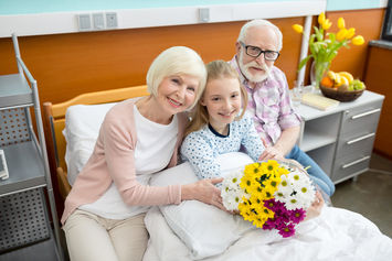 A smiling elderly couple sits beside a young girl holding a bouquet of flowers on a hospital bed. Hospital equipment and a table with fruit and flowers are in the background.